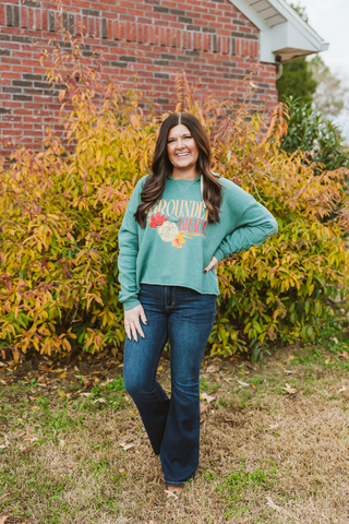 Brunette girl wearing grounded & grace long sleeve top with dark blue flare jeans in a grass field in front of a bush.