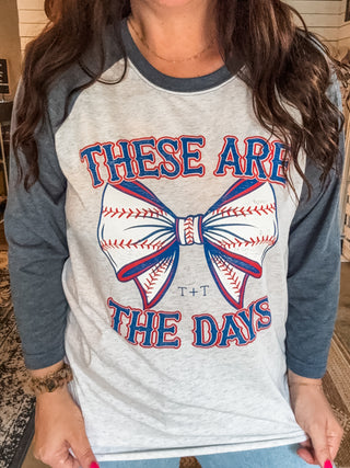 female wearing graphic tee in a baseball style with a neutral body and heather navy sleeves with print that reads "these are the days" in red and blue ink and a bow in the middle with baseball design in the bow