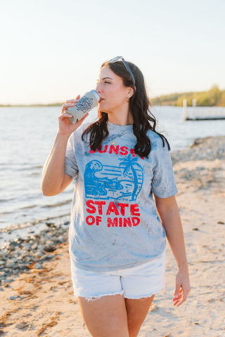 female model at a lake wearing shorts and a graphic tee that reads "sunset state of mind"