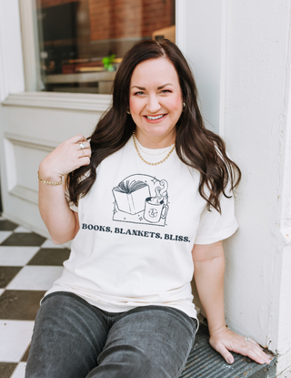 Woman wearing a white t-shirt with 'Books, Blankets, Bliss' design sitting on a step.
