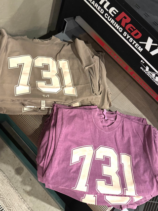 two graphic tees lying on a conveyer dryer reading 731 area code in cream color ink
