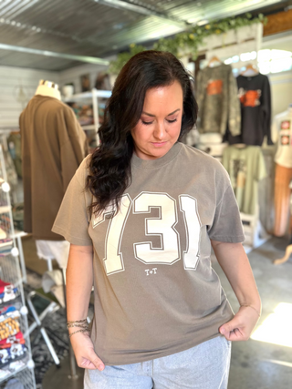 female wearing a taupe color graphic tee with a 731 design screen printed on the front in a cream ink with grey jeans