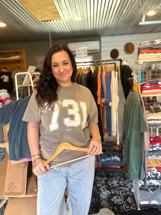 Crystal Tucker, founder of Tucked & Threaded, standing in her she-shed with hanging racks of clothes and hanger in her hand showcasing 731 graphic tee