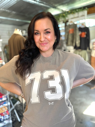 female wearing a taupe color short-sleeve graphic tee that has a cream color 731 design printed on the front