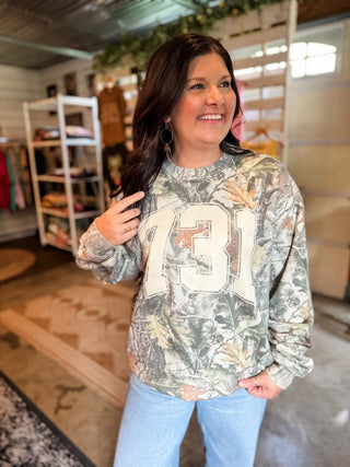 A woman wearing a camouflage long-sleeve sweatshirt with a large cream-colored '731' graphic on the front, paired with light blue jeans, standing inside a retail store.