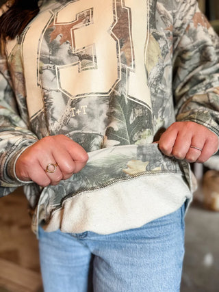 female model wearing blue jeans and tucked & threaded area code 731 sweatshirt in camo pattern and has 2 hands flipping up the bottom seam of the sweatshirt to show the fleece-lined inside