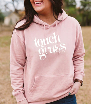 A woman wearing a dusty pink pullover hoodie with 'touch grass' printed in white across the chest, photographed outdoors in a blurred grassy field.