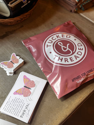 Pink packaging with 'Tucked & Threaded' logo, butterfly card, and small butterfly figurine on a wooden surface. | Monthly Graphic Tee Subscription for Women and Moms