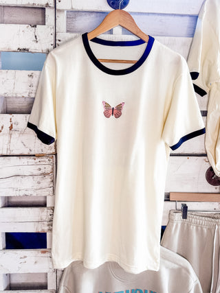 White t-shirt with a butterfly design on a wooden hanger against a rustic wooden wall in the she-shed