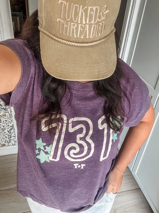 A person wearing a heathered purple graphic t-shirt with '731' and 'T-T' printed on the front, paired with a khaki baseball cap that reads 'TUCKED & THREADED'.