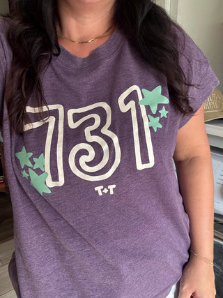 A person wearing a heather purple short-sleeve graphic T-shirt with large white outlined '731' text, mint green star accents, and small 'T+T' lettering on the front.