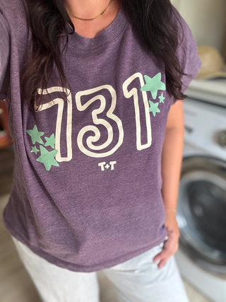 A person wearing a heathered purple graphic T-shirt with large cream outlined numbers '731', small green stars, and 'T+T' printed below, paired with light pants in a laundry room setting.