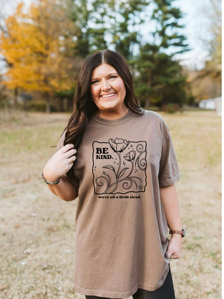 Woman wearing a brown t-shirt with 'Be Kind' graphic in an outdoor setting