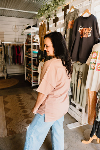 female model wearing graphic tee in nude color and blue jeans and she is showing the side split of the tee where the tee is longer in the back than the front