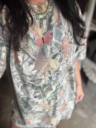 A close-up of a person wearing a short-sleeve camouflage-patterned top with a pink and gold butterfly applique at the center chest. The camouflage features shades of gray, green, brown, and beige.