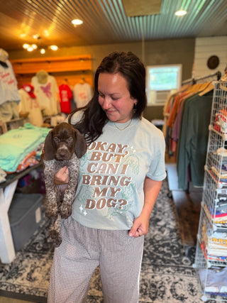 Woman holding a dog in a store with a shirt that says 'Okay, but can I bring my dog?'
