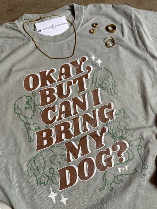 Gray t-shirt with 'Okay, but can I bring my dog?' text and dog illustrations on a flat surface.