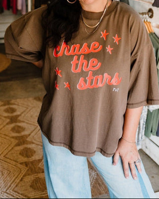 A woman wearing a brown oversized graphic t-shirt with 'Chase the stars' printed in orange, standing in a clothing boutique.