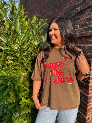A woman wearing a brown graphic t-shirt with red text reading 'Chase the stars' and small red star accents, standing outdoors next to a brick wall and green bushes.