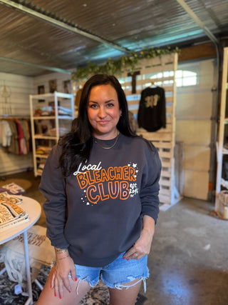 A woman wearing a dark gray crewneck sweatshirt with 'Local Bleacher Club' printed in orange and white on the front, paired with denim shorts, standing inside a retail store.