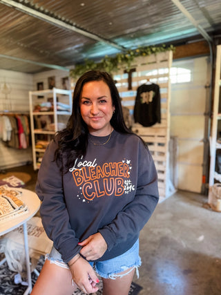 A woman wearing a dark gray crewneck sweatshirt with 'Local Bleacher Club' printed in orange and white on the front, standing inside a retail store.