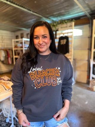 A woman with long dark hair stands in a retail store wearing a dark gray crewneck sweatshirt with 'Local Bleacher Club' printed in orange and white on the front, paired with light blue denim shorts.