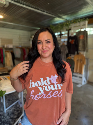 A woman with long dark hair stands inside a retail store wearing a rust-orange graphic t-shirt that reads 'hold your horses' in white and lavender script with a purple star. The background shows clothing racks and a store interior.