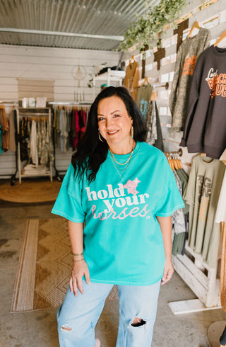 teal graphic tee reading "hold your horses" in pink and white ink worn by a female model in a she-shed with blue jeans
