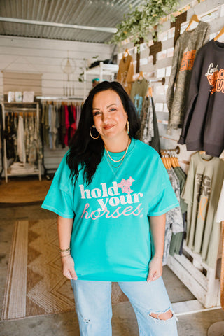 model wearing a graphic tee in a teal color that reads "hold your horses" in white and pink ink