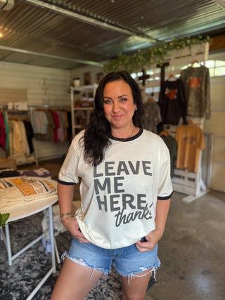 A woman posing inside a rustic clothing boutique, wearing a cream-colored graphic t-shirt with black lettering that reads 'LEAVE ME HERE thanks' and light-wash denim shorts.
