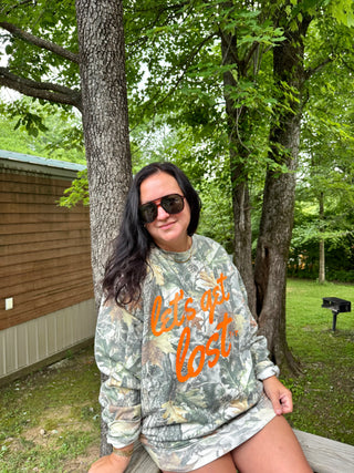 A woman wearing a camouflage sweatshirt with orange text reading 'Let's not get lost' and sunglasses, sitting outdoors in front of trees.