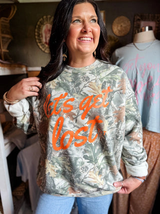 female model inside workspace of tucked & threaded wearing blue jeans and a "let's get lost" camo sweatshirt with the design printed in orange ink