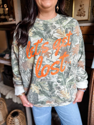 female model inside tucked & threaded she-shed standing in blue jeans and her graphic sweatshirt that reads "let's get lost" in orange ink on a camo print sweatshirt