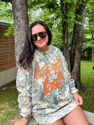A woman wearing a camouflage sweatshirt with orange text reading 'let's get lost' sits outdoors on a bench in front of trees and a cabin.
