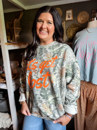 female model inside she-shed of tucked & threaded wearing blue jeans, her brunette hair down, and has on a "let's get lost" camo sweatshirt