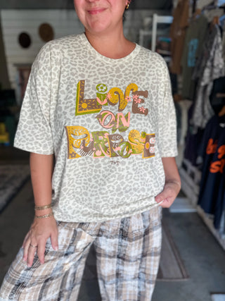 female model wearing a graphic tee in a solid leopard natural color with a graphic design printed on the front in plum, gold, coral, and green inks that reads "live on purpose" and tee is paired with plaid pants