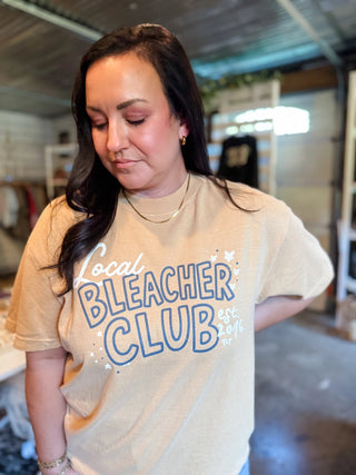 A woman wearing a beige graphic t-shirt with 'Local Bleacher Club' printed in blue and white, standing in a workshop or studio environment.