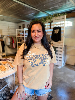 A woman with long dark hair stands inside a boutique shop wearing a beige graphic t-shirt that reads 'Local Bleacher Club' in blue and white, paired with denim shorts.