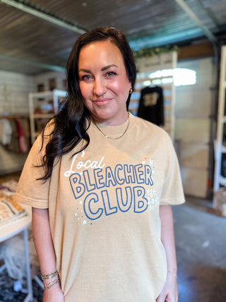 A woman with long dark hair stands indoors wearing a beige graphic T-shirt with the words 'Local Bleacher Club' printed in blue and white. The background shows a retail or workshop-like interior with shelves and equipment.