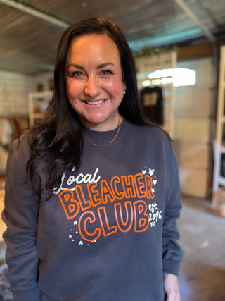 A smiling woman with long dark hair wearing a dark gray crewneck sweatshirt with a colorful graphic that reads 'Local Bleacher Club est. 2016' in orange, white, and black, set against a blurred garage-like background.
