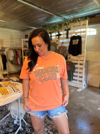 A woman wearing a bright orange graphic t-shirt that reads 'Local Bleacher Club' in white and gray, paired with denim shorts, standing inside a retail store.