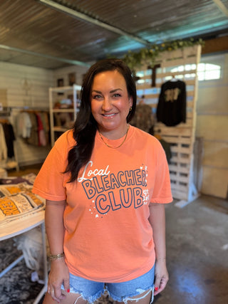 A woman smiling while wearing a bright coral orange graphic t-shirt that reads 'Local Bleacher Club' in stylized lettering, paired with light blue denim shorts inside what appears to be a retail store.