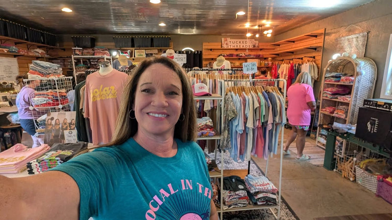 A woman taking a selfie inside a retail store, wearing a teal t-shirt with pink text that reads 'SOCIAL IN THE SUNSHINE"