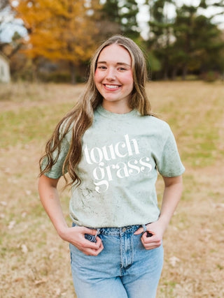 A smiling young woman stands outdoors in a grassy field wearing a light sage green t-shirt with the words 'touch grass' printed in white script, paired with light-wash blue jeans.