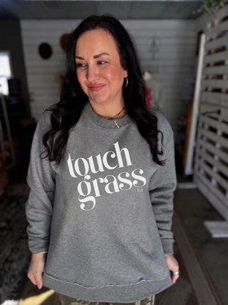 A woman with long dark hair wearing a heather gray crewneck sweatshirt with 'touch grass' printed in white across the chest, standing in an indoor setting.