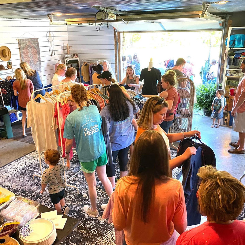A busy boutique interior with multiple people browsing clothing racks and standing near an open garage-style door. Shoppers are seen holding garments, with shirts, dresses, and other apparel displayed throughout the store.