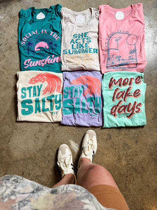Six folded graphic T-shirts arranged in two rows on a concrete floor, with a person's legs and sneakers visible at the bottom. The shirts feature colorful retro designs and text including 'Social in the Sunshine,' 'She acts like summer,' 'Stay Salty,' 'Stay Salty AF,' and 'more lake days.