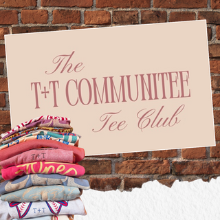 Stack of colorful t-shirts with a sign reading 'The T+T Communittee Tee Club' against a brick wall | Tucked and Threaded