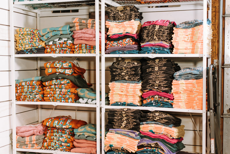 Stacks of folded colorful garments and textiles arranged on white metal shelving units in a storage or retail environment.