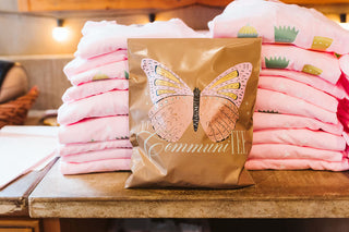 A brown paper gift bag with a butterfly graphic and the word 'communi' stands in front of stacks of folded pink t-shirts on a wooden table.